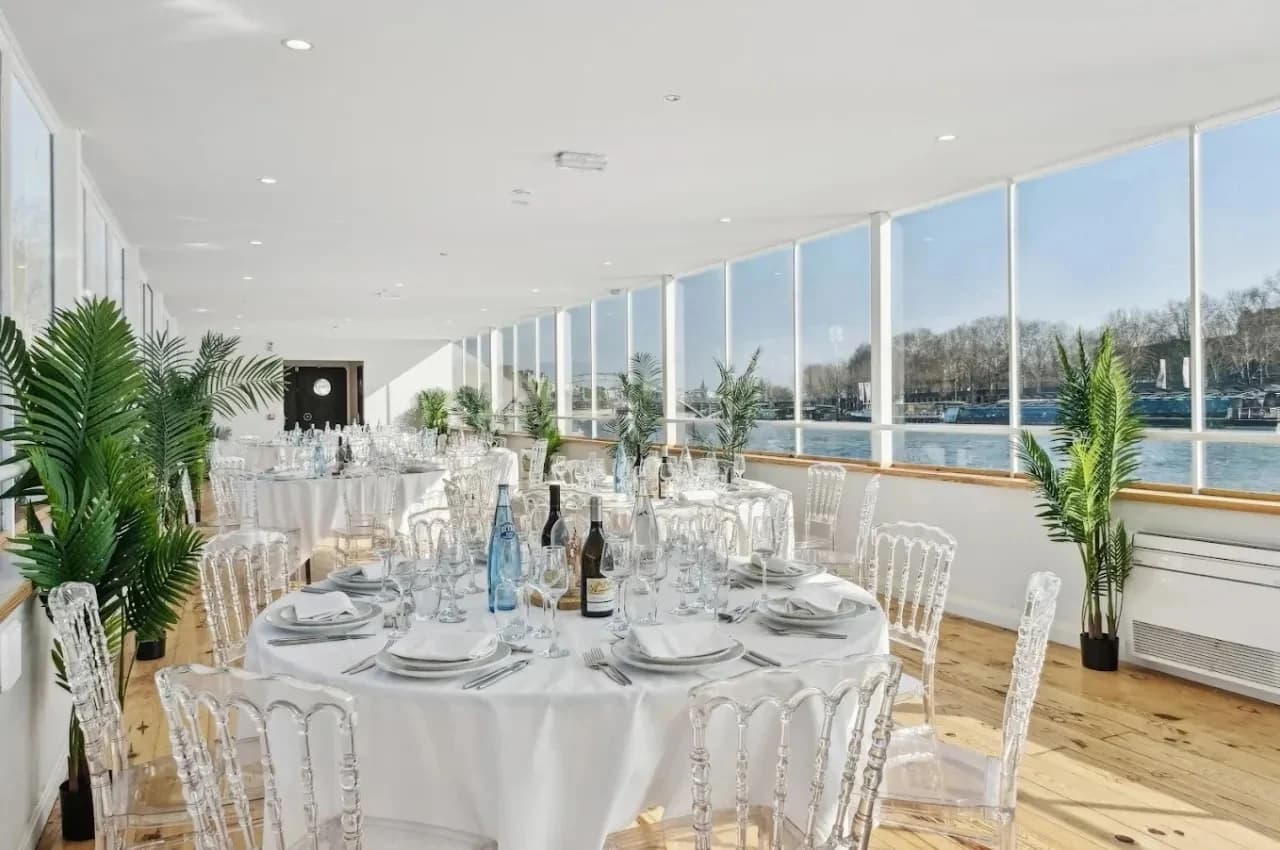Reception room on a barge with white tables, clear chairs and a wall of windows over the river.