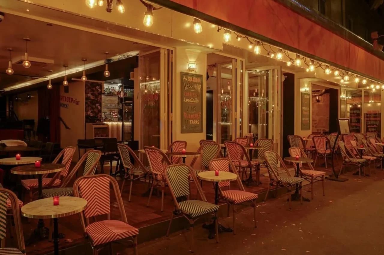 Street terrace of a venue at dusk with striped chairs, string lights and a chalkboard menu.