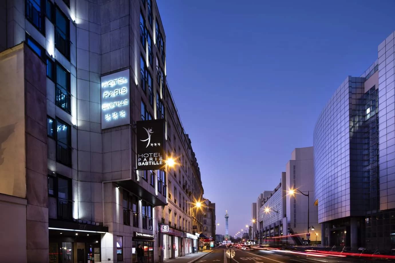 Exterior of Hôtel Paris Bastille at dusk, with its illuminated hotel sign on a busy street in Paris