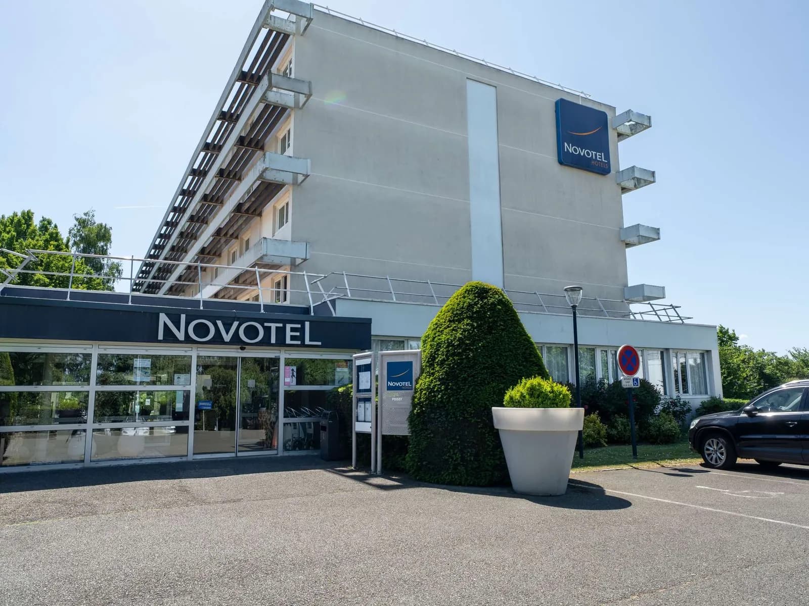 Front of the Novotel Poissy Orgeval hotel with its entrance canopy, car park and blue brand sign