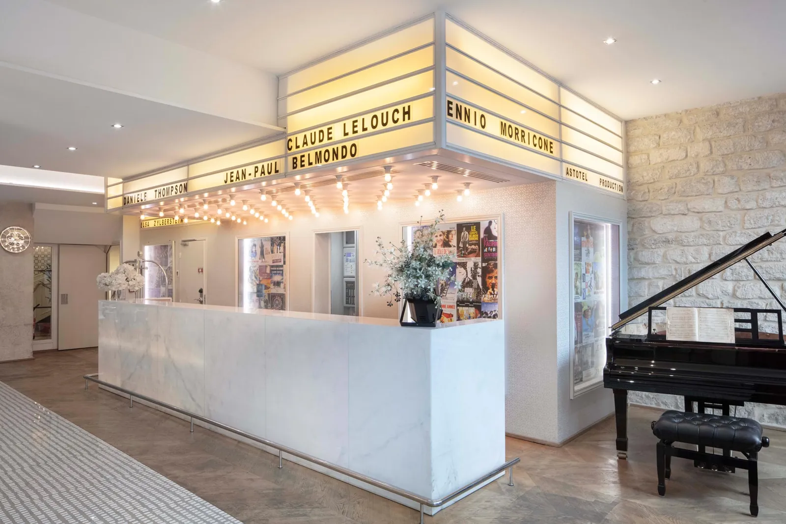 Comptoir de réception sous un fronton lumineux de noms de films, avec un piano à queue noir à côté, au Le 123 Sébastopol