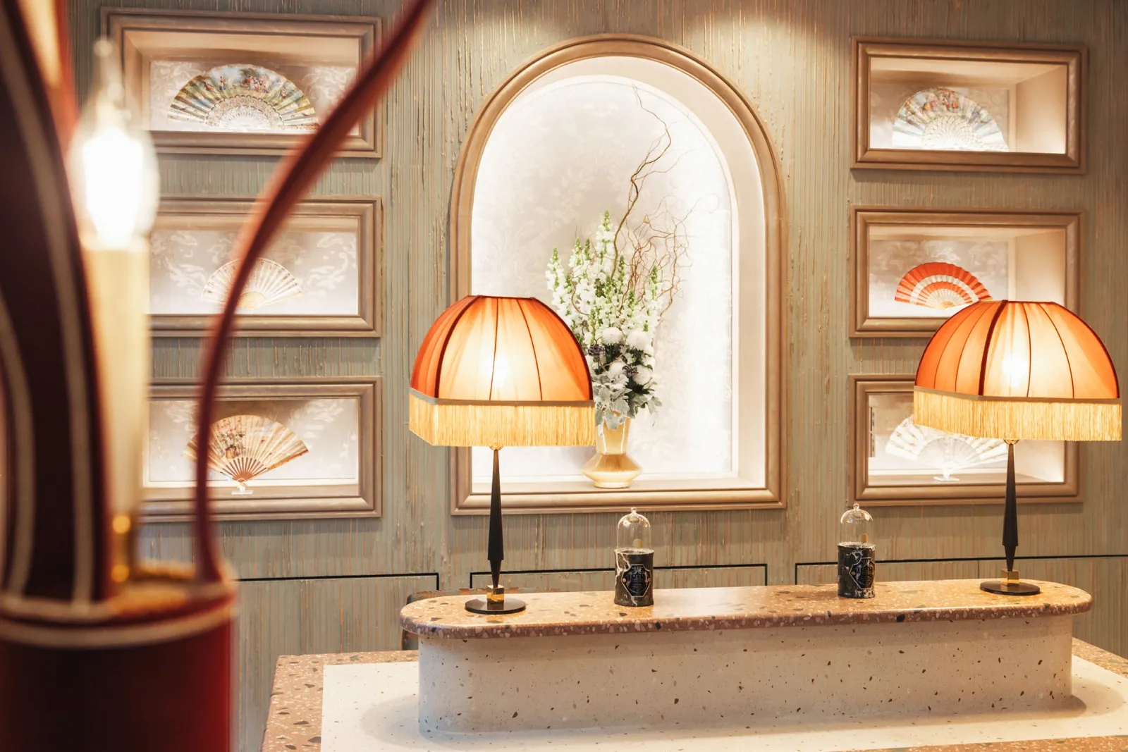 Espace d'accueil de la Maison Barrière Vendôme avec lampes à franges, niche cintrée et éventails encadrés