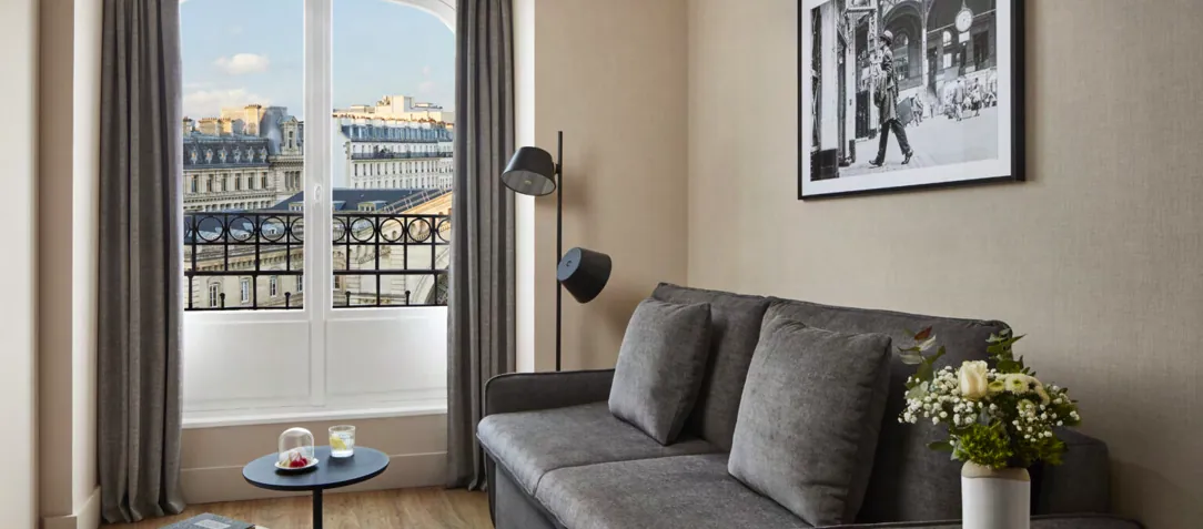 Chambre du NH Paris Gare de l'Est avec canapé gris et fenêtre donnant sur les toits et balcons parisiens
