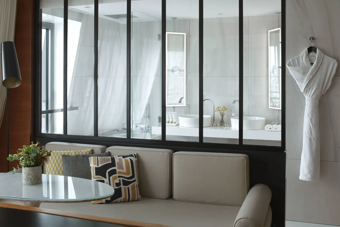 Suite seating area with grey sofa and bathroom glimpsed through glass at Renaissance Paris République Hotel