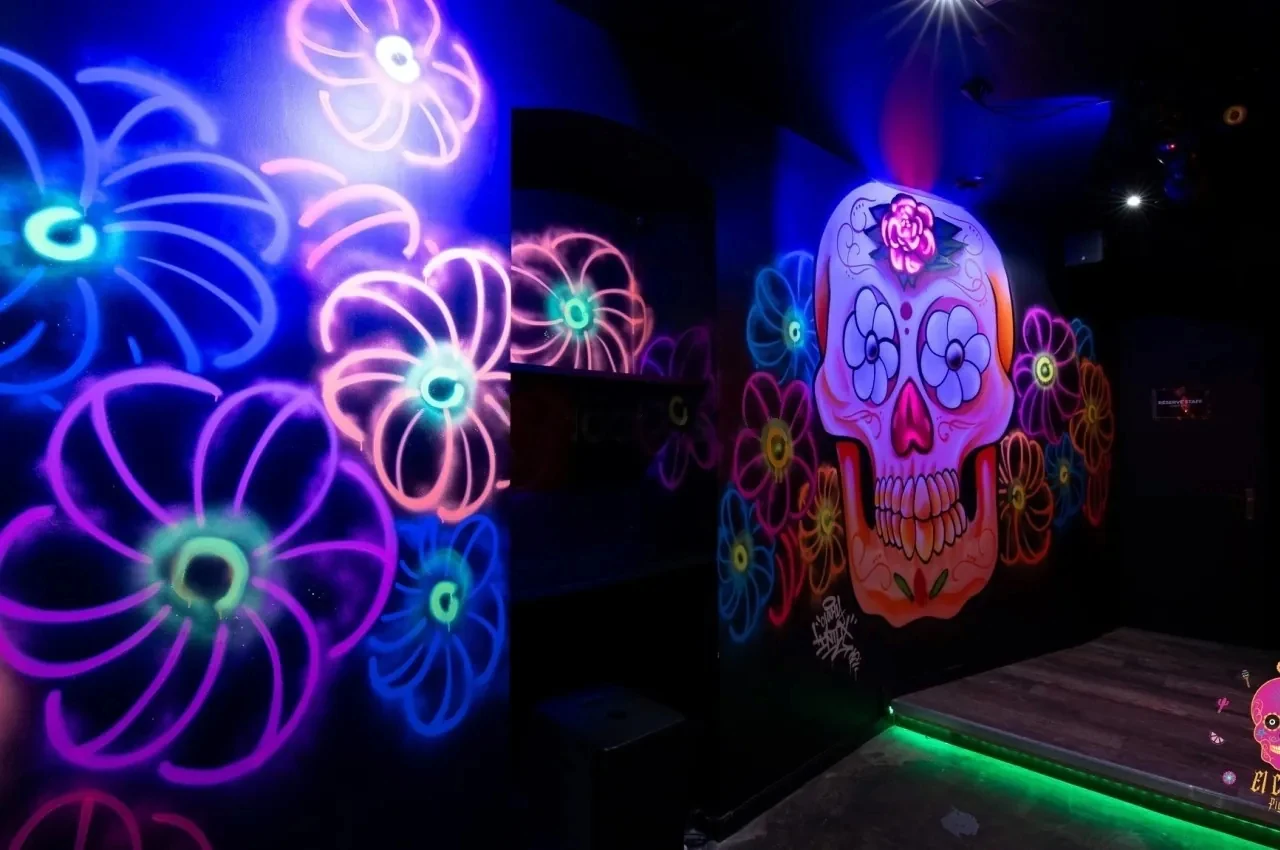 Dark room with neon flower murals and a large skull painting lit in blue and green.