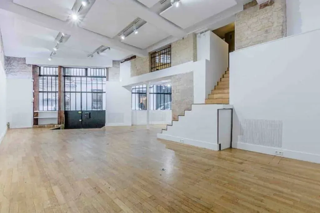 Grande salle aux fenêtres à cadres noirs, parquet et escalier vers la mezzanine