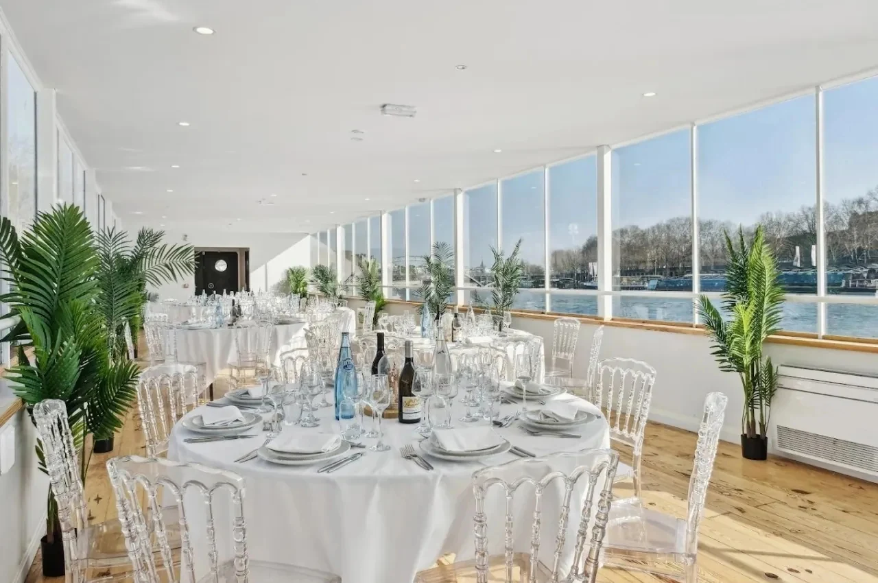 Reception room on a barge with white tables, clear chairs and a wall of windows over the river.