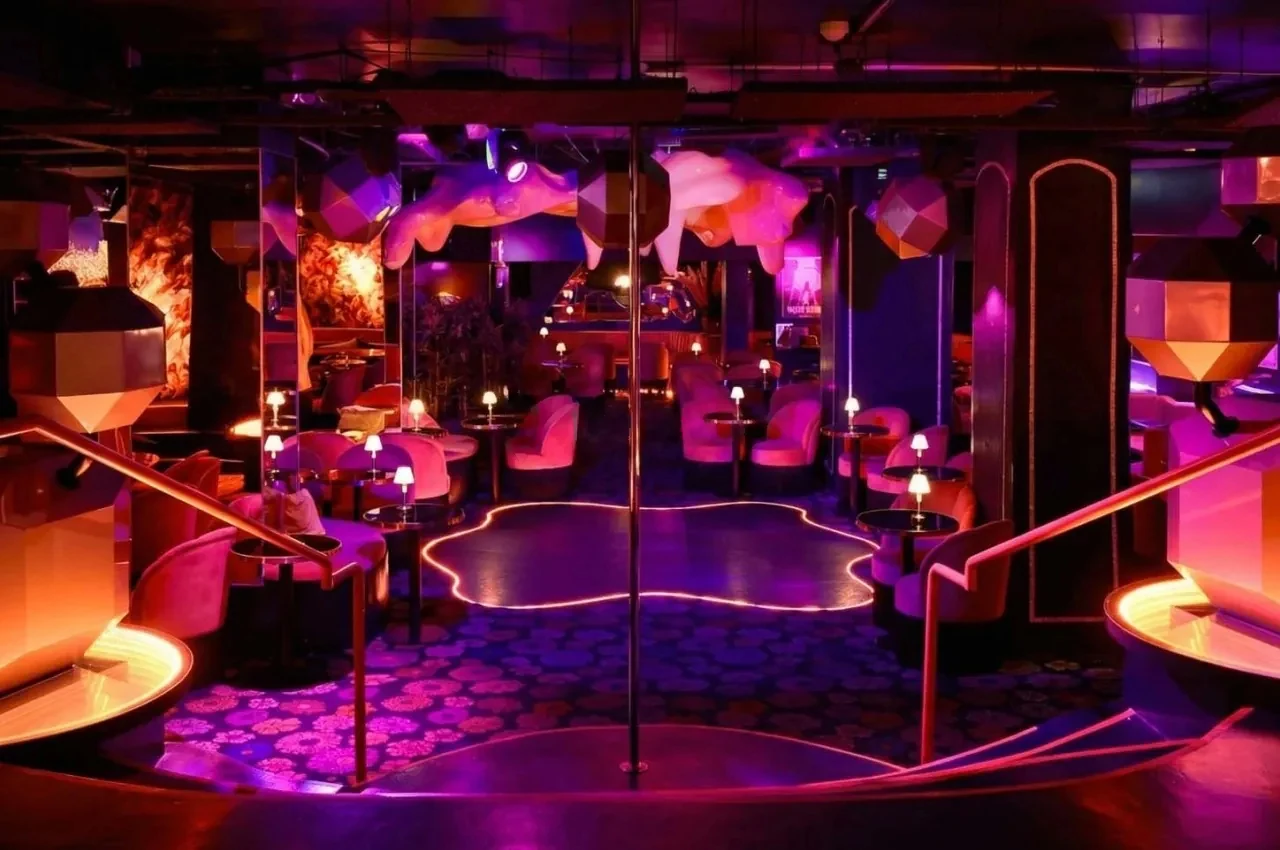 Club room with pink and violet lighting, round lounge seats and a central pole.