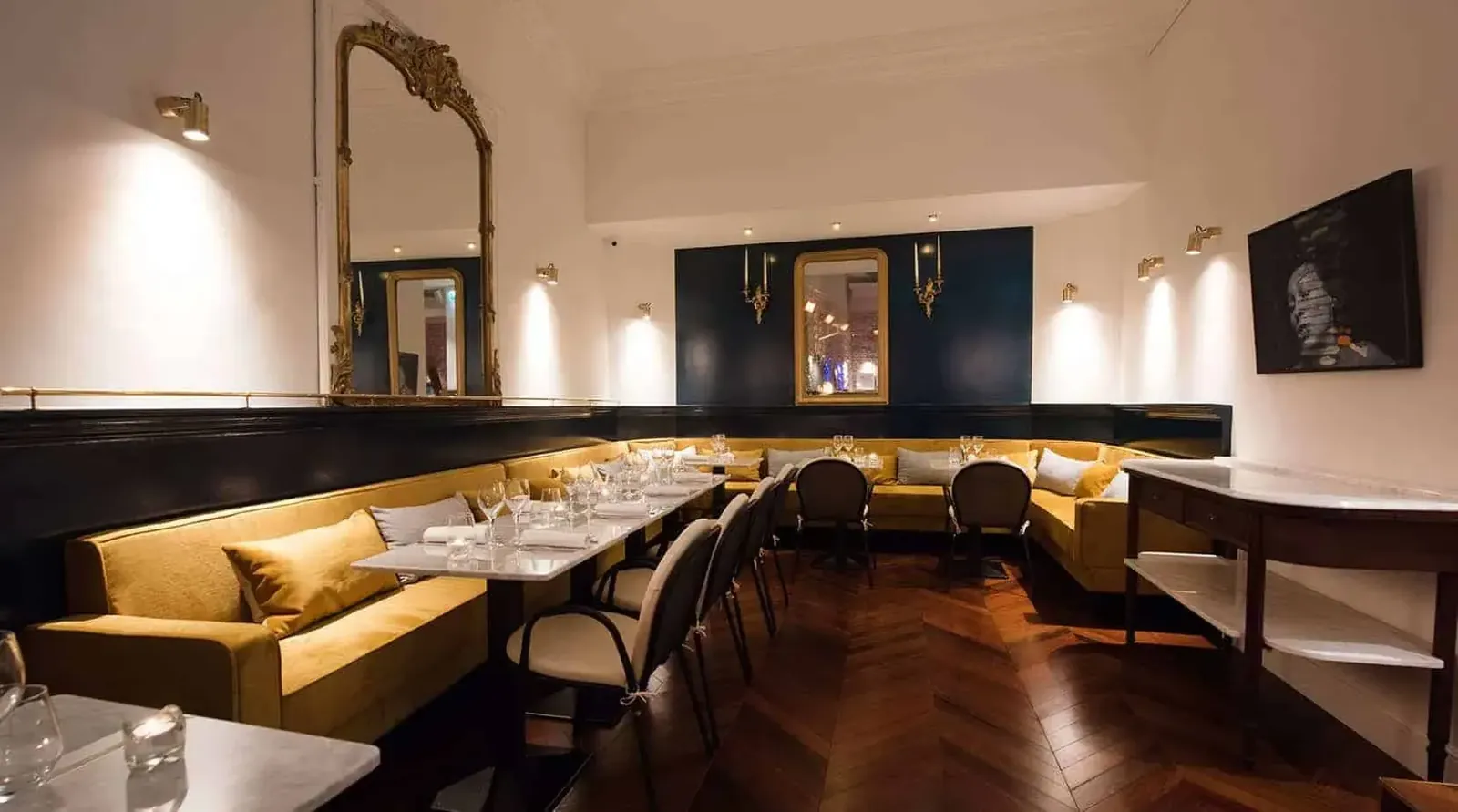 Dining area with yellow velvet banquettes, a gilded mirror and a dark parquet floor