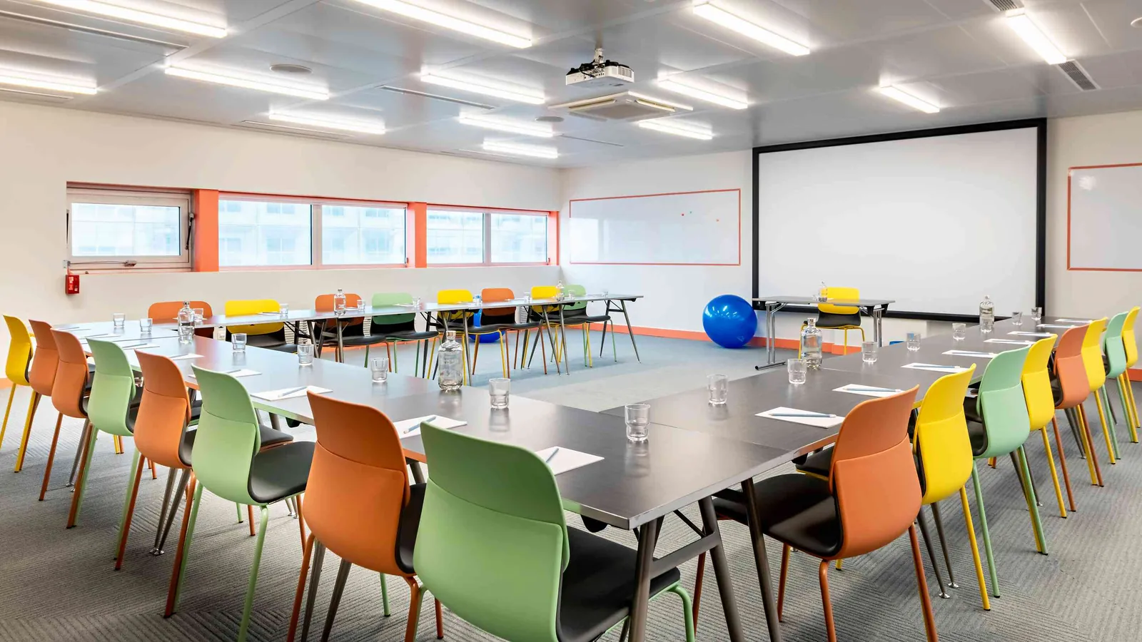Salle plénière du Pavillon Ouest avec chaises orange, vertes et jaunes autour de tables longues