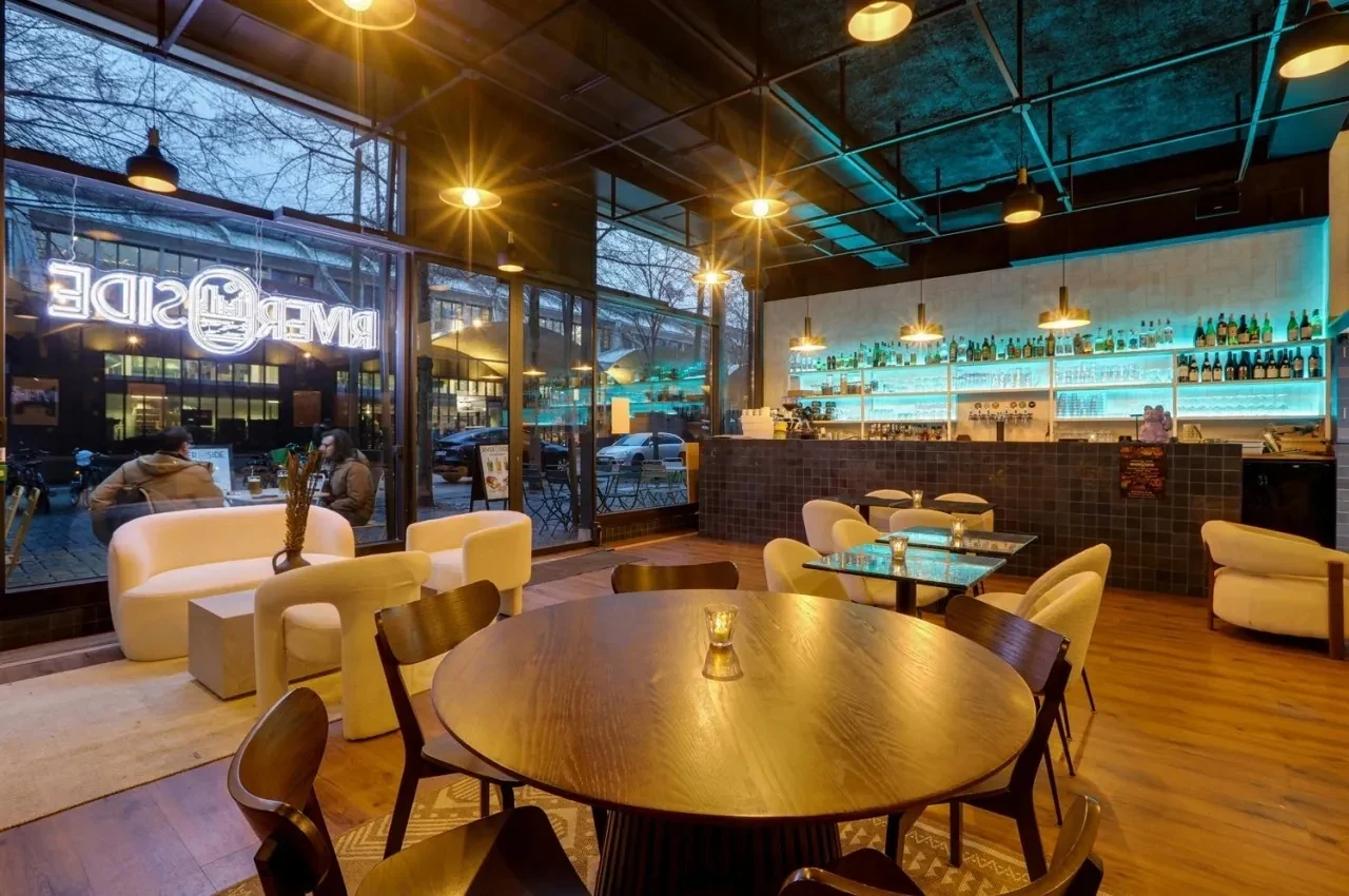 Lounge bar with a neon RIVERSIDE sign, white armchairs and a round dining table.